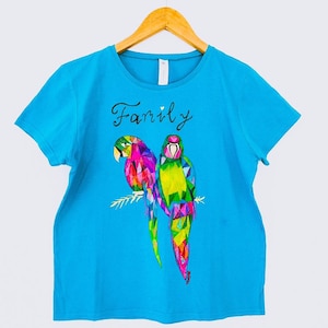 Puede incluir: Una camiseta azul turquesa de manga corta presenta un diseño geométrico colorido de dos loros posados en una rama. La palabra "Family" está escrita en cursiva negra sobre los loros.