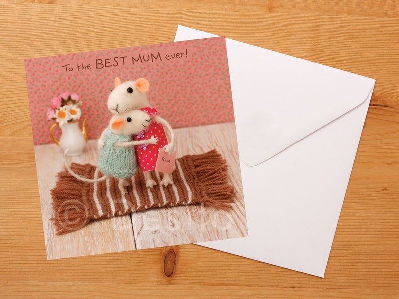 May include: A greeting card with a pink background featuring two white felt mice hugging on a brown and white striped rug. The card reads "To the BEST MUM ever!"