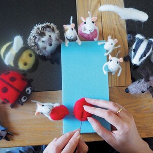 May include: A variety of needle felted animal figurines, including a bee, hedgehog, ladybug, mice, a badger, and a dog. The figurines are arranged in a circle around a blue piece of foam. A person's hands are shown working on a red felt ball.