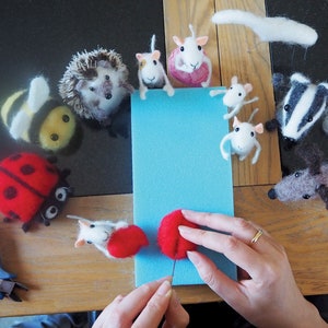 May include: A variety of needle felted animal figurines, including a bee, hedgehog, ladybug, mice, a badger, and a dog. The figurines are arranged in a circle around a blue piece of foam. A person's hands are shown working on a red felt ball with a needle.