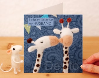 May include: A birthday card with a blue background featuring two white felt giraffes with brown spots and red ears. The card has the text "Birthday kisses for my HUSBAND" on the front.