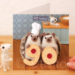May include: A birthday card featuring two felt hedgehogs wearing chef hats holding a biscuit with the number 90 on it. The card says "90 today!" and has a blue and white patterned background.