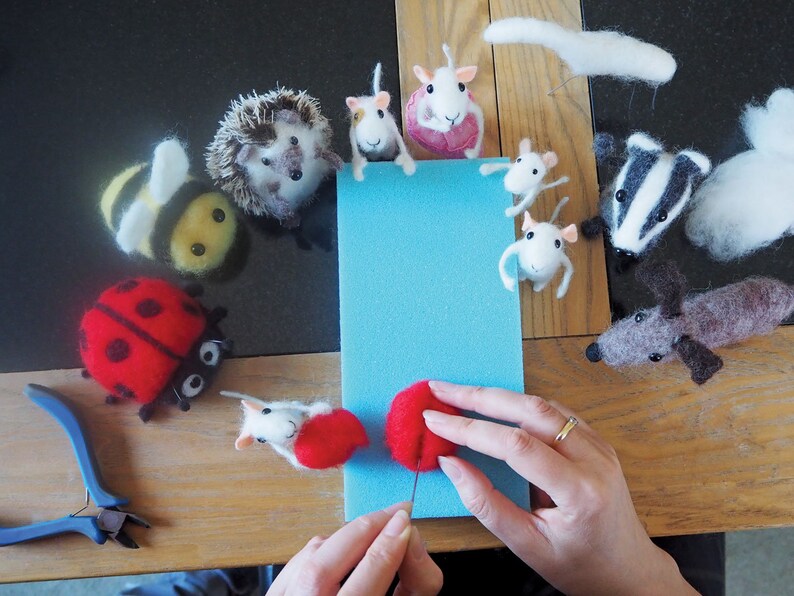 May include: A variety of needle felted animal figurines, including a bee, hedgehog, ladybug, mice, a badger, and a dog. The figurines are arranged in a circle around a blue piece of foam. A person's hands are shown working on a red felt ball.