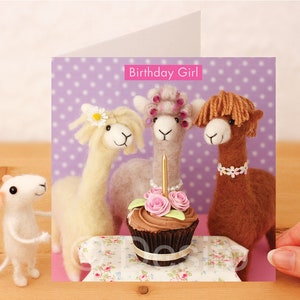 May include: A birthday card featuring three alpaca figurines with a cupcake and a single lit candle. The card has a purple background with white polka dots and the text "Birthday Girl!" in pink.