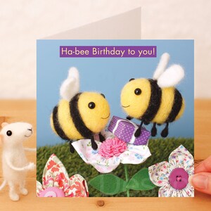 May include: A birthday card featuring two felt bumblebees with black and yellow stripes, holding a purple gift box. The card has a blue background and the text "Ha-bee Birthday to you!" in purple.