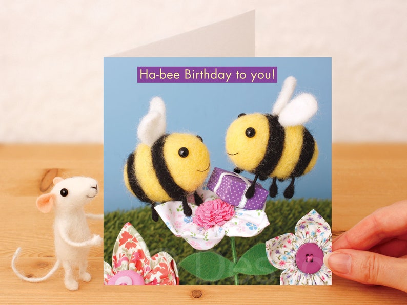 May include: A birthday card featuring two felt bumblebees with black and yellow stripes, holding a purple gift box. The card has a blue background and the text "Ha-bee Birthday to you!" in purple.