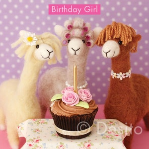 May include: Three fluffy alpaca figurines with a chocolate cupcake topped with pink frosting and a gold candle. The alpaca in the center has pink hair rollers and a pearl necklace. The alpaca on the right has a brown collar with white flowers. The card reads "Birthday Girl".