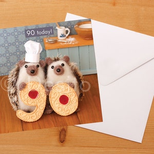 May include: Two brown and white hedgehogs wearing chef hats hold up two large biscuits shaped like the number 90. The biscuits have red jelly centers. The text "90 today!" is in the top left corner of the card.