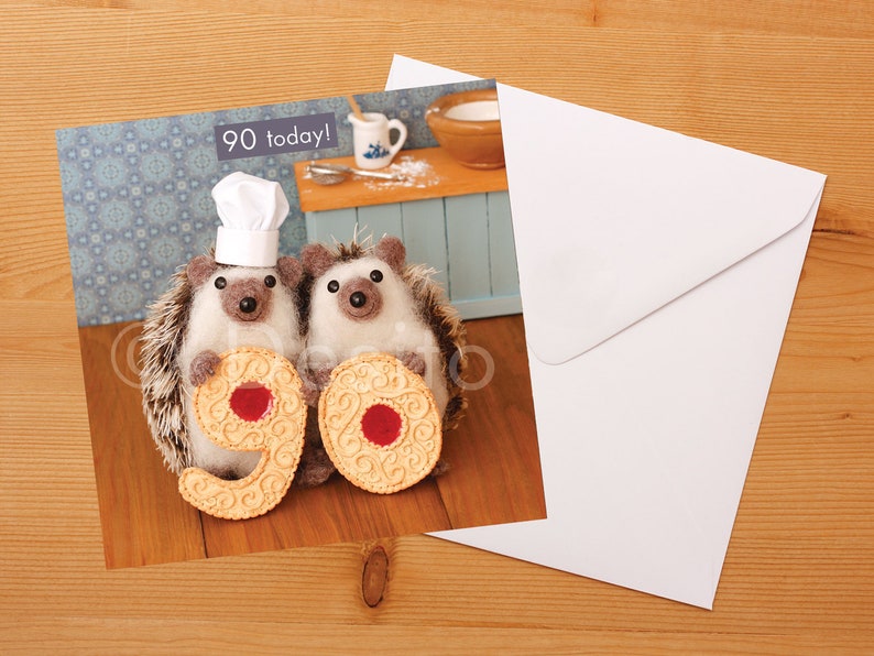 May include: Two brown and white hedgehogs wearing chef hats hold up two large biscuits shaped like the number 90. The biscuits have red jelly centers. The text "90 today!" is in the top left corner of the card.