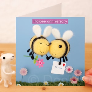 May include: A greeting card with a blue background featuring two felt bees with white wings and black stripes. The bees are holding a note that says "To you" with a red heart. The card says "Ha-bee anniversary" in pink letters.