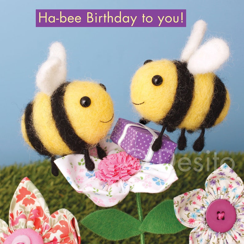 May include: Two yellow and black bumblebees with white wings are perched on a white flower with pink accents. One bee is holding a purple gift box with white polka dots. The card reads "Ha-bee Birthday to you!"