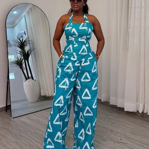 Puede incluir: Mono turquesa con cuello halter y estampado de triángulos blancos. El mono tiene un pantalón de pernera ancha y un corpiño ajustado. El atuendo se usa con gafas de sol y accesorios dorados. El mono es un atuendo elegante y cómodo.