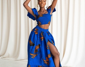 May include: A woman wears a blue crop top and a long blue skirt with an orange print. The skirt has a high slit on the right side.