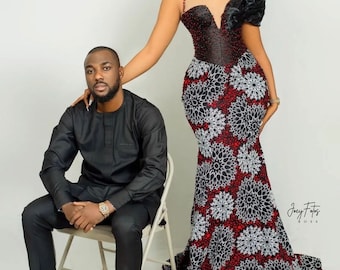 Puede incluir: Una mujer con un vestido formal y un hombre con un traje negro posan para un retrato. El vestido presenta un corpiño negro con una falda estampada floral roja y blanca. El hombre está sentado en una silla, vistiendo un traje negro y mocasines verdes.