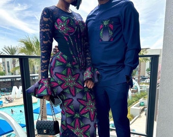 May include: A couple wearing matching blue and green patterned outfits. The woman is wearing a long-sleeved dress with a lace overlay and the man is wearing a long-sleeved shirt and pants. They are both smiling and looking at the camera.