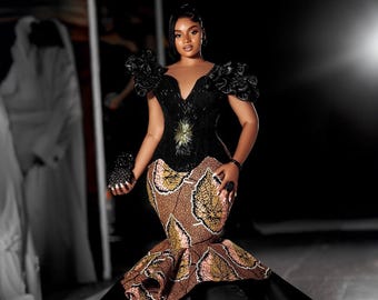 May include: A black and brown mermaid-style gown with a fitted bodice and a flowing skirt. The bodice features a black sequined design and a large gold embellishment. The skirt is made of a brown fabric with a leaf pattern.