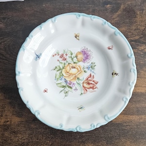 Könnte beinhalten: Weißer Keramikteller mit hellblauem, gewelltem Rand. Der Teller zeigt einen Blumenstrauß mit gelben Rosen, lila Blumen und roten Tulpen. Kleine Insekten sind um die Blumen verstreut.