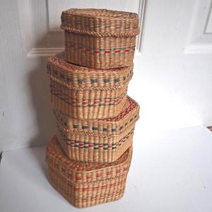 May include: A stack of four hexagonal woven baskets with lids. Each basket features natural tan reed construction with horizontal bands of red and dark green woven accents. The baskets are arranged vertically, decreasing in size toward the top.