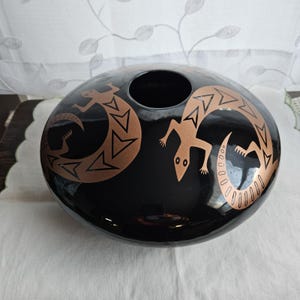 Puede incluir: Un jarrón de cerámica negro con forma redonda y una gran abertura. Dos diseños de lagartos de color cobre están pintados en la superficie. Los lagartos están estilizados con patrones geométricos.
