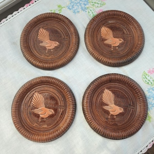 May include: Set of four round, wooden coasters. Each coaster features a carved bird design with the words "NEW ZEALAND" in a circular pattern. The coasters are a warm brown color and have a ridged edge.