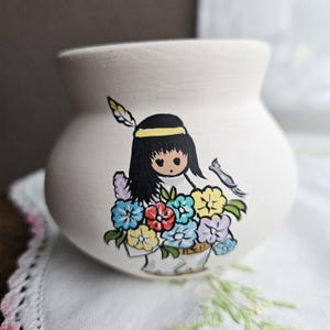 Puede incluir: Un pequeño jarrón de cerámica blanquecina de forma redondeada. El jarrón está pintado a mano con un diseño de una niña nativa americana, una cesta de flores coloridas y un pájaro. La niña tiene pelo negro, una diadema amarilla y una pluma.