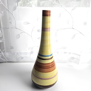 May include: A tall, slender ceramic vase with a tapered neck and a rounded base. The surface features horizontal stripes in shades of yellow, brown, light blue, and soft pink, creating a layered, multi-colored pattern across the entire body.