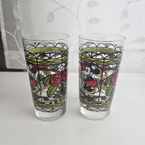 Może przedstawiać: Dwie przezroczyste szklanki z kolorowym nadrukiem w stylu witrażu przedstawiającym myszkę Minnie. Wz&oacute;r zawiera czarne kontury, czerwone r&oacute;że, zielone liście i białe kwiaty ułożone w powtarzający się wz&oacute;r łuk&oacute;w na zewnętrznej powierzchni.