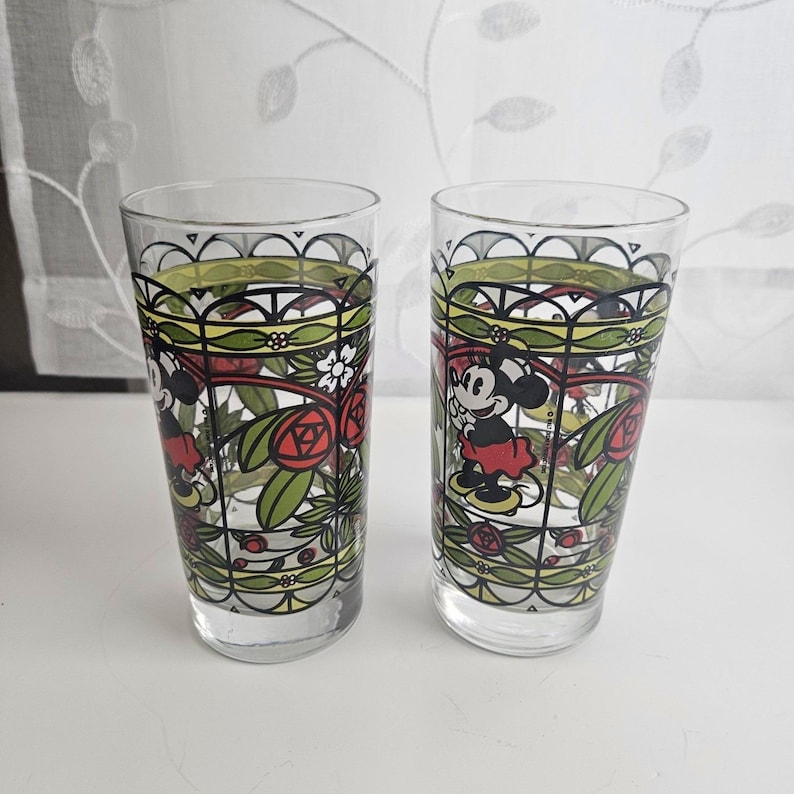 Może przedstawiać: Dwie przezroczyste szklanki z kolorowym nadrukiem w stylu witrażu przedstawiającym myszkę Minnie. Wz&oacute;r zawiera czarne kontury, czerwone r&oacute;że, zielone liście i białe kwiaty ułożone w powtarzający się wz&oacute;r łuk&oacute;w na zewnętrznej powierzchni.