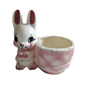Könnte beinhalten: Ein weißer Keramik-Pflanztopf in Form eines Kaninchens, das einen rosafarbenen und weißen Tonnen-Pflanztopf hält. Das Kaninchen hat rosa Ohren und schwarze Augen.