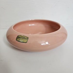 May include: A round, light pink ceramic bowl with a smooth, glossy finish. A gold-colored rectangular sticker is affixed to the side, featuring the black text Haeger Floral, Macomb, IL, Made in U.S.A. The bowl has a wide, curved rim.