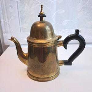 Può includere: Una teiera in ottone caratterizzata da un coperchio alto e bombato con un puntale, un beccuccio curvo e un manico nero. Il corpo in metallo presenta una finitura lucida con segni visibili di usura e patina, di forma cilindrica e con coperchio.