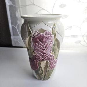 Może przedstawiać: Biały ceramiczny wazon z wypukłym, rzeźbionym wzorem fioletowego kwiatu irysa z zielonymi liśćmi. Wazon ma rozszerzany brzeg i wycięcia w górnej części, ukazujące szczegółowy, teksturowany relief kwiatowy na przedniej powierzchni naczynia.