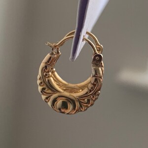 Könnte beinhalten: Goldene Creolen mit einem detaillierten, verzierten Design. Die Creolen sind dick und haben eine abgerundete Form.