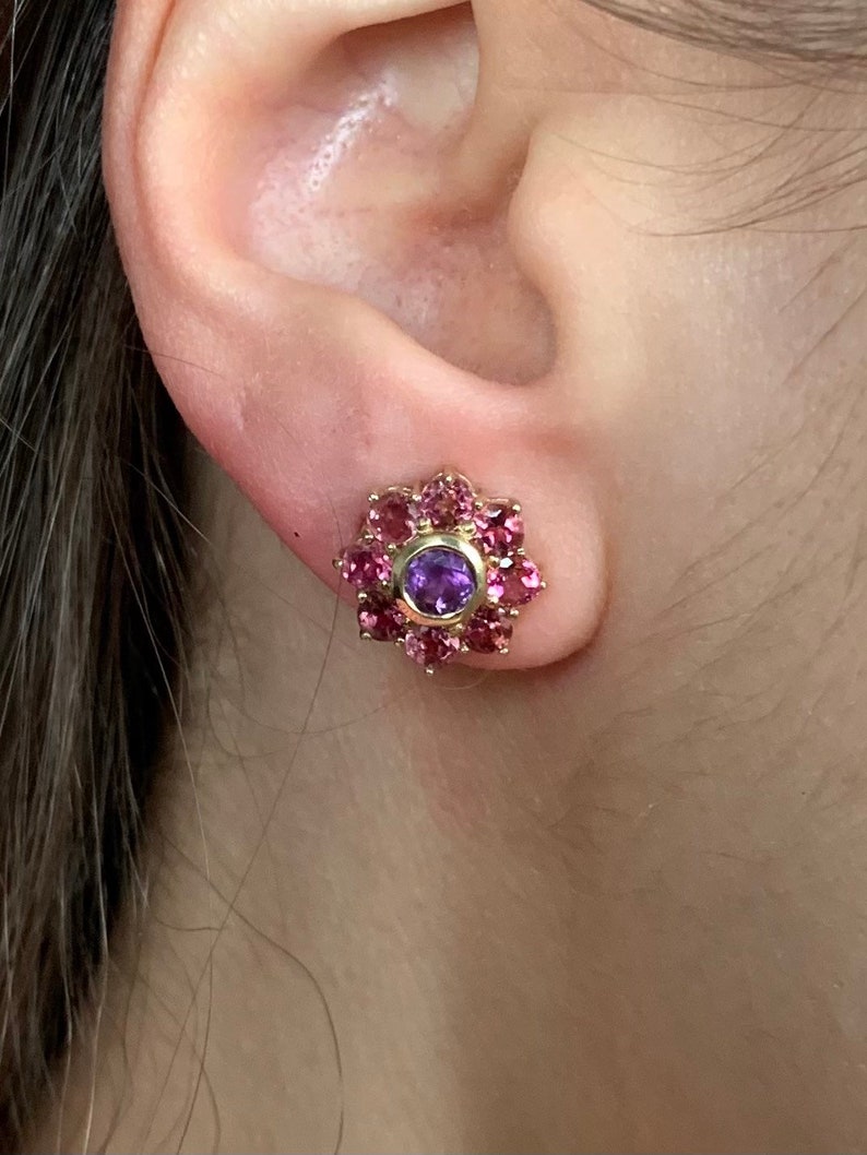 K&ouml;nnte beinhalten: Ein Paar goldene Ohrringe mit einem violetten Edelstein, umgeben von rosa Edelsteinen.