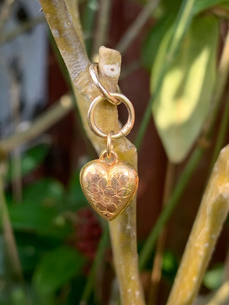 May include: A gold heart charm with a floral design hangs from a gold chain. The charm is attached to a gold jump ring.