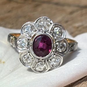 Peut inclure: Une bague vintage avec un motif floral. La bague présente un rubis central ovale, entouré d'un halo de diamants ronds et clairs. Le corps de la bague est un mélange de tons or et argent, avec des détails complexes.