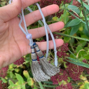 Pode incluir: Um cordão prateado com duas borlas prateadas e uma pequena conta roxa. O cordão é segurado em uma mão contra um fundo de folhagem verde.
