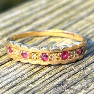 Könnte beinhalten: Ein goldener Ring mit einer Reihe kleiner, runder, roter Edelsteine. Der Ring hat einen strukturierten, dekorativen Rand und liegt auf einer Holzoberfläche. Die Edelsteine sind gleichmäßig angeordnet.