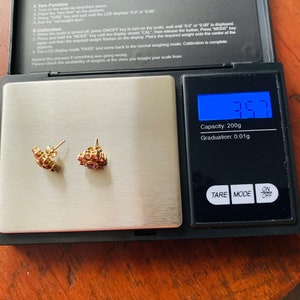 K&ouml;nnte beinhalten: Nahaufnahme einer kleinen digitalen Waage mit einem Paar goldenen Ohrringen mit rosa Steinen auf der Plattform. Die Waage zeigt 3,57 Gramm an. Die Waage hat eine Kapazit&auml;t von 200 Gramm und eine Ablesegenauigkeit von 0,01 Gramm.
