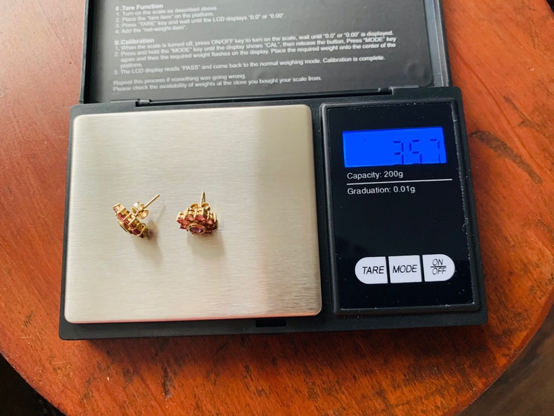 K&ouml;nnte beinhalten: Nahaufnahme einer kleinen digitalen Waage mit einem Paar goldenen Ohrringen mit rosa Steinen auf der Plattform. Die Waage zeigt 3,57 Gramm an. Die Waage hat eine Kapazit&auml;t von 200 Gramm und eine Ablesegenauigkeit von 0,01 Gramm.