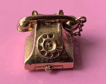 May include: A gold-toned rotary phone charm with a chain attached. The phone has a detailed design and a realistic look.