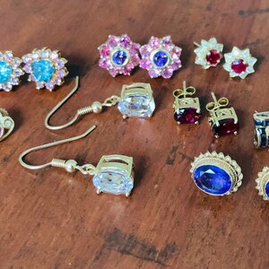 K&ouml;nnte beinhalten: Eine Auswahl an Ohrringen mit verschiedenen farbigen Edelsteinen, darunter blau, lila, rot und wei&szlig;. Die Ohrringe sind aus Gold und in verschiedenen Stilen erh&auml;ltlich, darunter Ohrstecker, H&auml;ngeohrringe und Ohrringe mit Anh&auml;nger.
