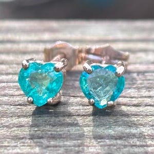 Puede incluir: Un par de pendientes de botón en forma de corazón. Cada pendiente presenta una vibrante piedra preciosa azul claro engastada en metal plateado. Las piedras preciosas están talladas en forma de corazón. Los pendientes se exhiben sobre una superficie de madera.
