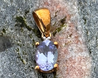 Pode incluir: Um pendente dourado com uma pedra preciosa ovalada de cor violeta clara. O pendente tem um design simples com uma argola dourada e quatro garras segurando a pedra. A pedra preciosa tem cerca de 1,3 cm de comprimento.