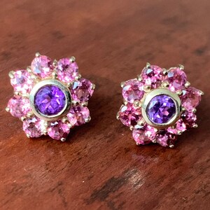 K&ouml;nnte beinhalten: Ein Paar goldene Ohrringe mit einem violetten Edelstein, der von rosa Edelsteinen in Bl&uuml;tenform umgeben ist.