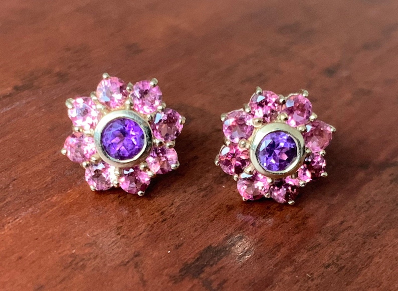 K&ouml;nnte beinhalten: Ein Paar goldene Ohrringe mit einem violetten Edelstein, der von rosa Edelsteinen in Bl&uuml;tenform umgeben ist.