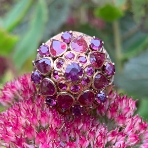 May include: A gold ring with a cluster of purple and pink gemstones. The ring is set with a variety of sizes and shapes of stones, creating a unique and eye-catching design.