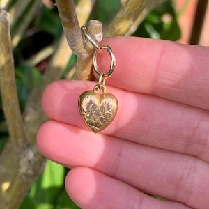 May include: A gold heart charm with a floral design. The charm is attached to a gold jump ring.