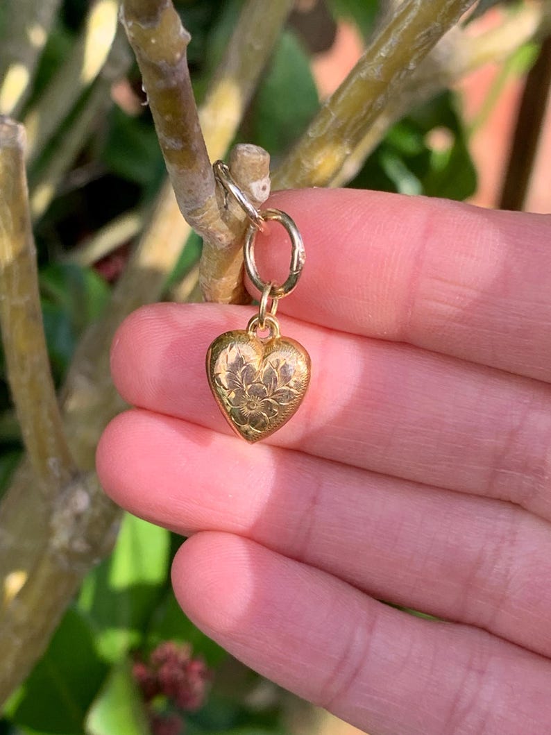 May include: A gold heart charm with a floral design. The charm is attached to a gold jump ring.