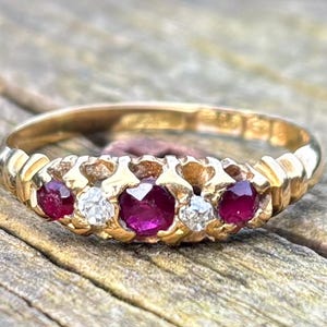 May include: A gold ring featuring five gemstones: two rubies and three diamonds. The rubies are a deep red, and the diamonds are clear. The ring is set on a wooden surface.